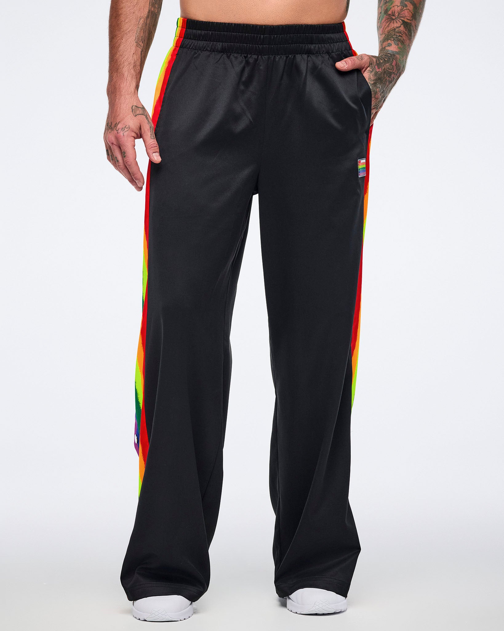 Male model wearing black track pants with rainbow side stripes and white sneakers, standing forward with his left hand in his pocket against a white background.