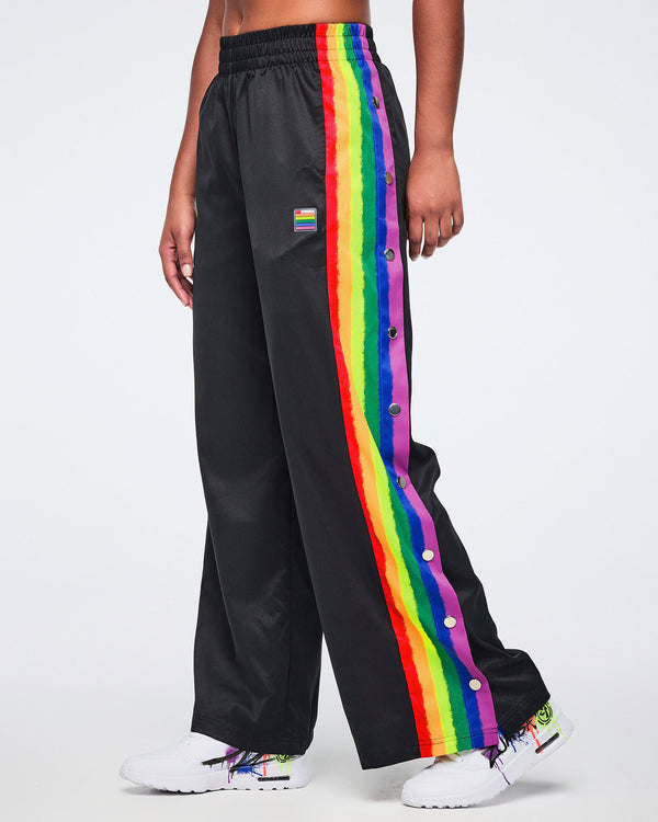 Female model wearing black track pants with rainbow side stripes and white sneakers, standing against a white background.