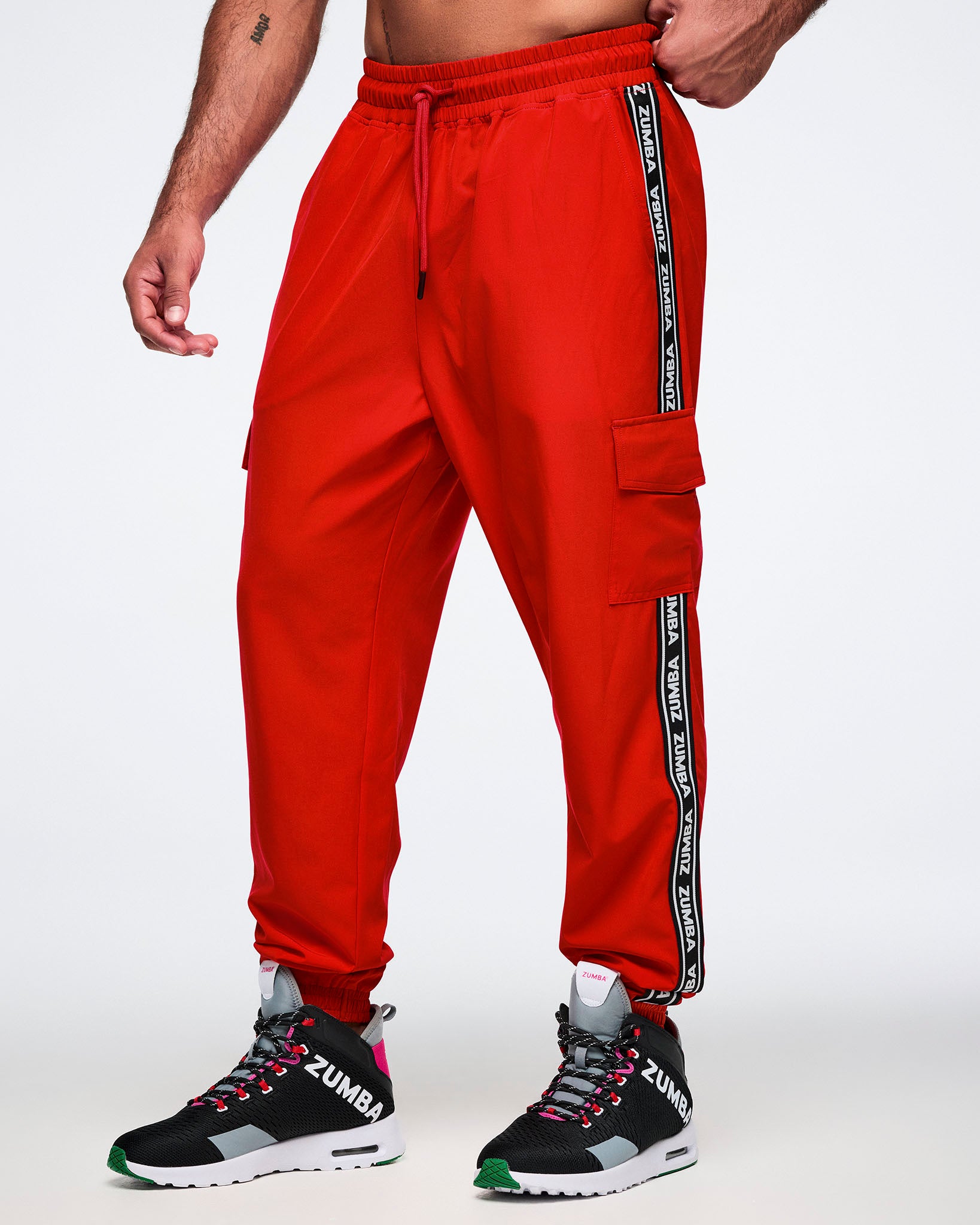 Male model wearing red cargo pants with black and white stripes saying 'Zumba' in white, paired with black and pink shoes on a white background.