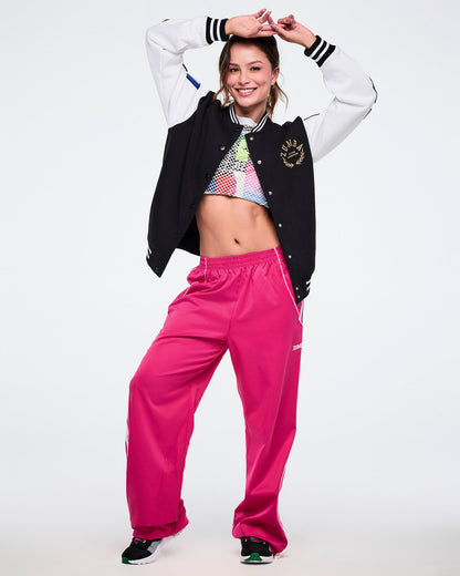 Female model with arms above her head, dancing, while wearing a black and white varsity jacket, colorful crop top, and pink pants on a white background.