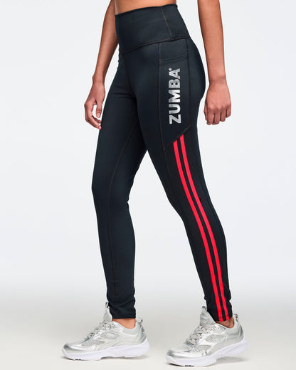 Black high-waisted leggings with silver vertical Zumba logo on the hip and bright red double side stripes. Female model wears fitted full-length leggings with a wide waistband and silver athletic sneakers, shown in a side view against a light gray background.