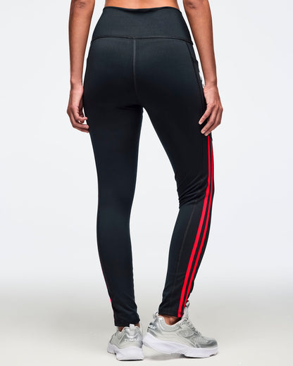 Black high-waisted leggings shown from the back with a sleek fitted silhouette, side pockets, and double coral pink side stripes. Female model is wearing the full-length leggings with silver athletic sneakers.