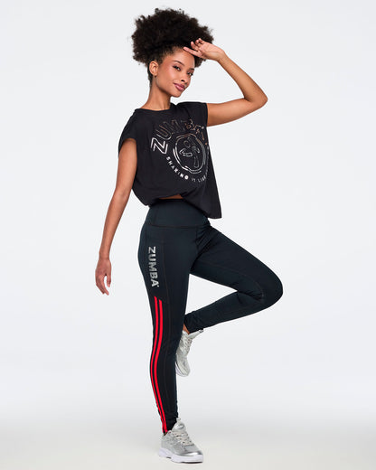 Black high-waisted leggings with silver vertical Zumba logo and bright red double side stripes. Female model wears fitted full-length leggings with a wide waistband, a black cropped Zumba graphic top, and silver athletic sneakers, posing in a full-body side view against a light gray background.