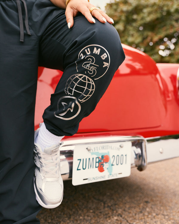 Black Zumba jogger pants with a silver “Zumba 25 Years” graphic down the leg. Female model wears the relaxed-fit black joggers with white crew socks, silver athletic sneakers, posing with one knee raised against a red vintage car.
