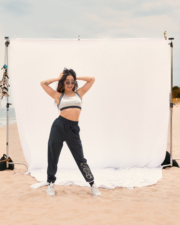 Black cargo jogger pants with a silver Zumba graphic on the lower leg. Female model wears the black joggers with a metallic silver sports bra, white sneakers, standing on sand in front of a white backdrop at the beach.