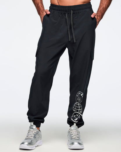 Black jogger pants with an elastic drawstring waistband, cargo pockets, and a silver Zumba 25 Years globe graphic on the lower leg. Male model is wearing the tapered joggers with silver athletic sneakers.