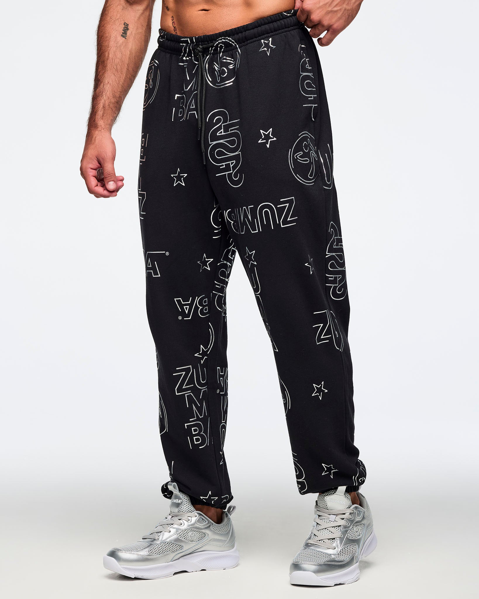 Black jogger sweatpants with an allover silver Zumba logo, icon, and star print, elastic drawstring waistband, and cuffed ankles. Male model is wearing the relaxed-fit joggers with silver athletic sneakers in a side view.