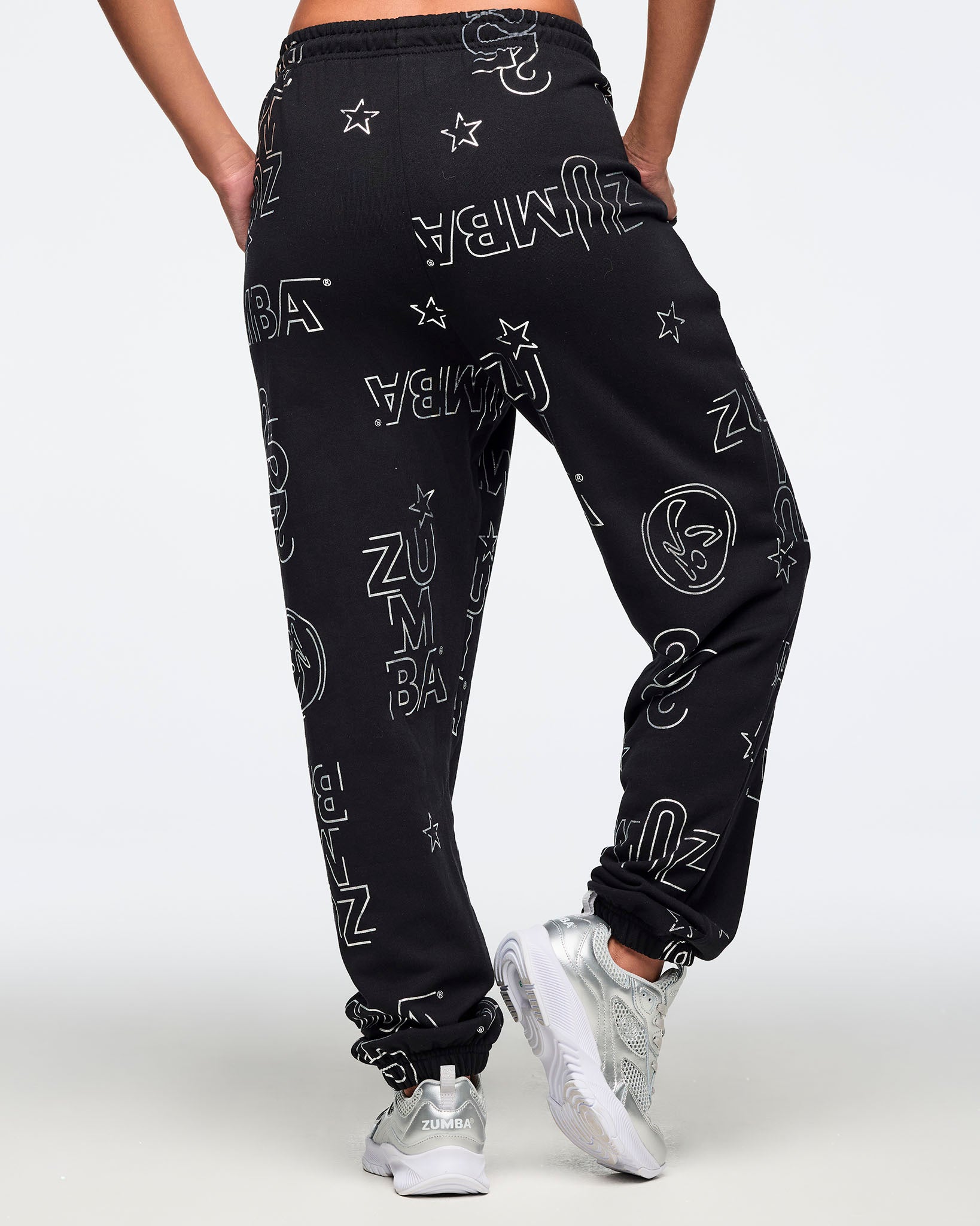 Black jogger sweatpants shown from the back with an allover silver Zumba logo, icon, and star print and cuffed ankles. Female model is wearing the relaxed-fit joggers with silver athletic sneakers.