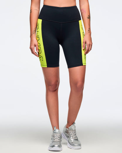 Black high-waisted biker shorts with neon yellow side panels and oversized black Zumba text. Female model is wearing the fitted above-the-knee shorts with silver athletic sneakers.