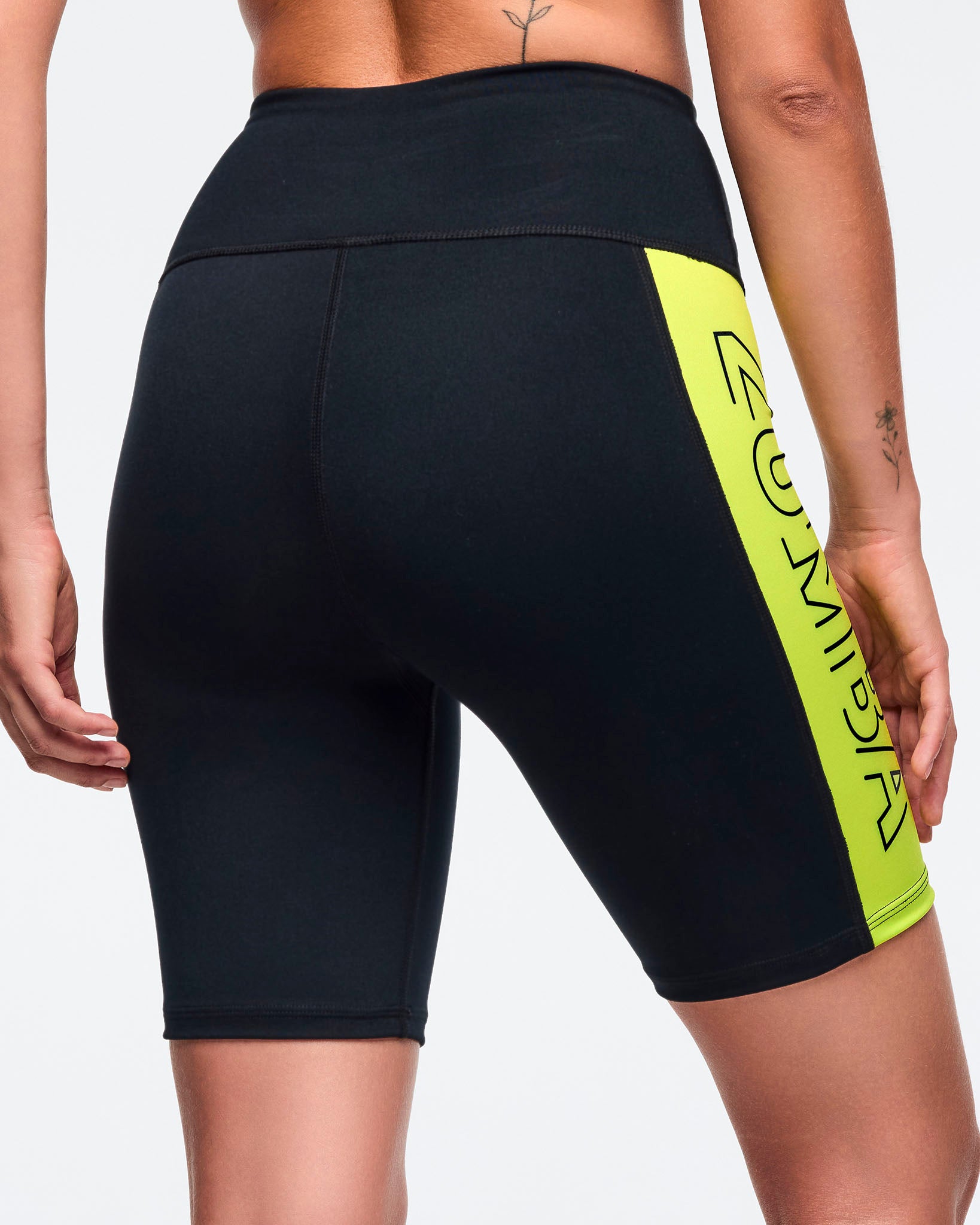 Black high-waisted biker shorts with a neon yellow side panel and partial vertical ZUMBA logo. Female model wears fitted mid-thigh performance shorts with a wide waistband, shown from the back to highlight the smooth black rear and side contrast panel against a light gray background.