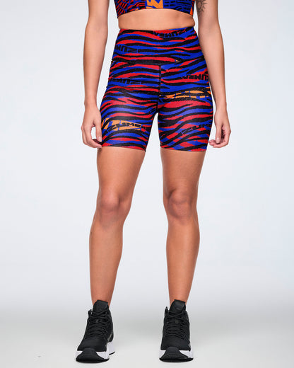 Female model wearing high-waisted biker shorts in a bright blue, red, black, and orange abstract zebra-print pattern, paired with a matching crop top and black high-top sneakers.