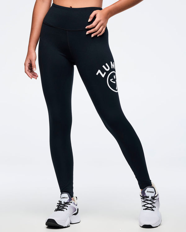 A female model wearing black high-waisted Zumba leggings with Zumba text and Zumbito logo, both in white, on the left thigh, paired with white and black athletic sneakers, shown from a side profile.