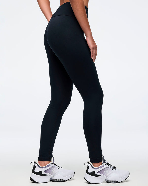 Back view of a female model wearing black high-waisted Zumba leggings, paired with white and black athletic sneakers.