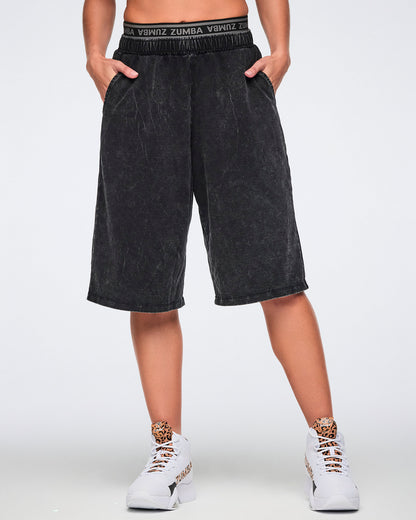 Black long pants with an oversized fit, side pockets, elastic Zumba logo waistband, and below-the-knee length, shown from the front on a female model wearing white high-top Zumba sneakers with cheetah-print tongue details.