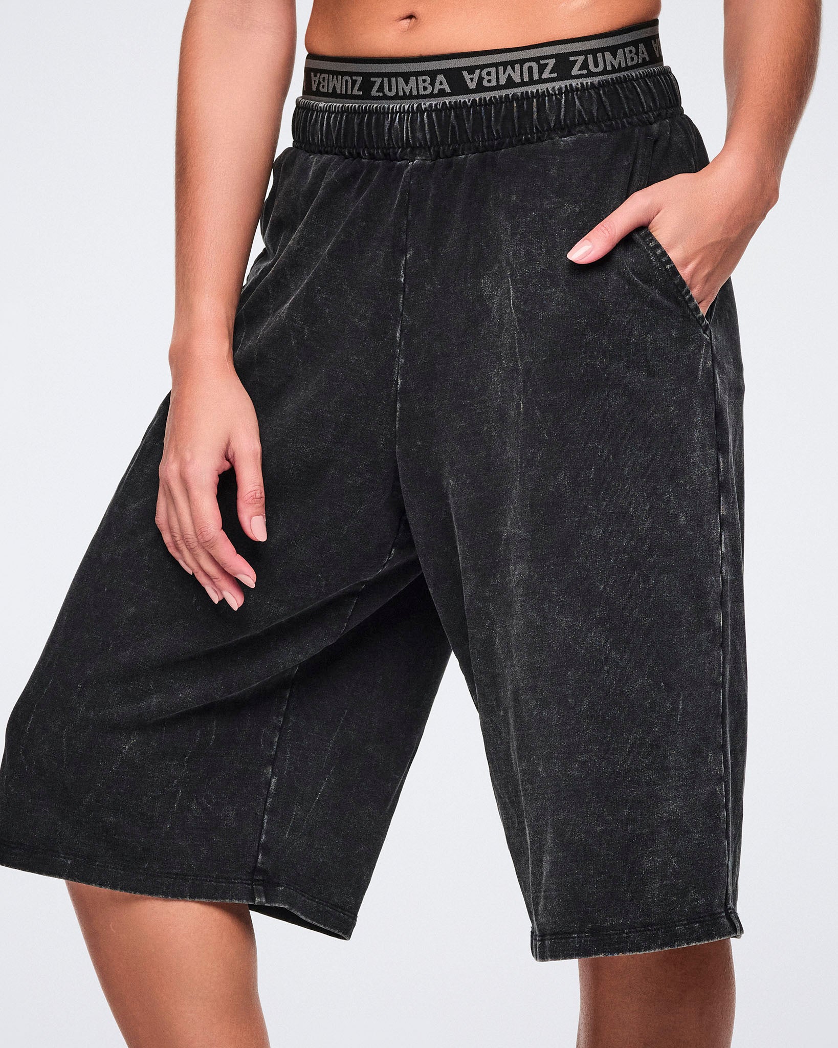 Close-up of black long shorts with an oversized fit to the knees, side pockets and elastic Zumba logo waistband shown on a female model.
