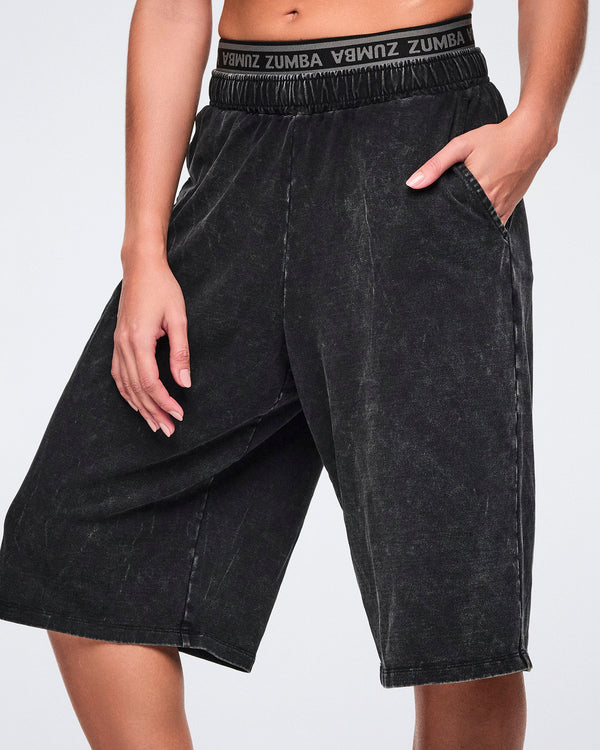 Close-up of black long shorts with an oversized fit to the knees, side pockets and elastic Zumba logo waistband shown on a female model.