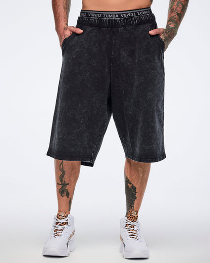 Black long pants with an oversized fit, side pockets, elastic Zumba logo waistband, and below-the-knee length, shown from the front on a male model wearing white high-top Zumba sneakers with black and cheetah-print details.