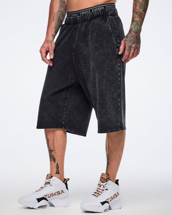 Black long shorts with an oversized fit, side pockets, and a black elastic Zumba logo waistband, shown on a male model wearing white high-top Zumba sneakers with black and cheetah-print details.