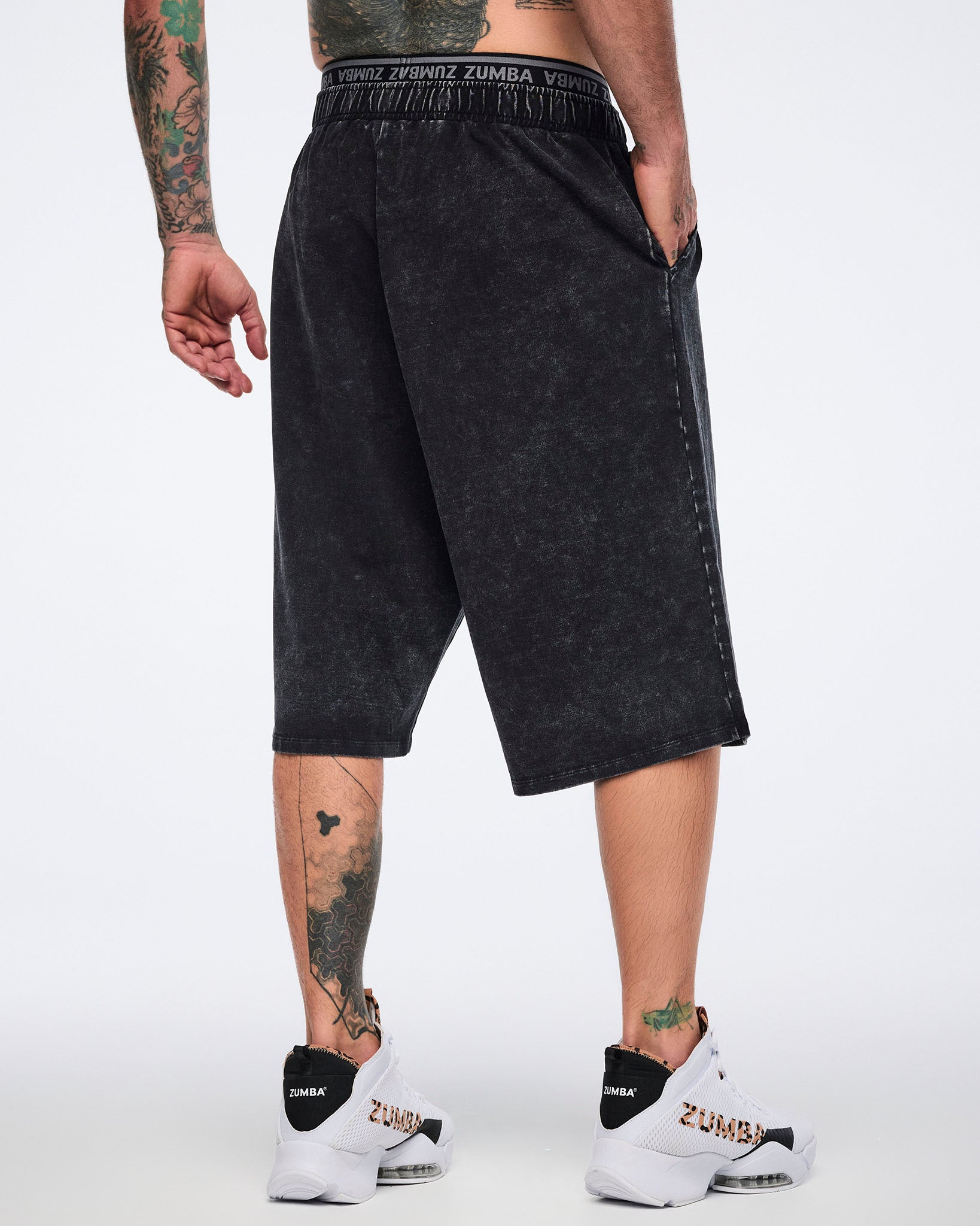 Black long pants with an oversized fit, elastic Zumba logo waistband, side pockets, and below-the-knee length, shown from the back on a male model wearing white high-top Zumba sneakers with black heel panels and cheetah-print details.