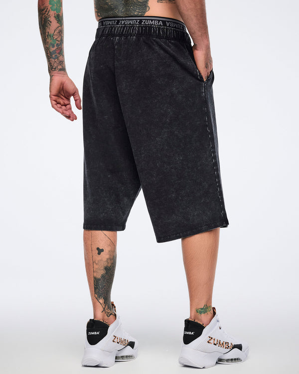 Black long pants with an oversized fit, elastic Zumba logo waistband, side pockets, and below-the-knee length, shown from the back on a male model wearing white high-top Zumba sneakers with black heel panels and cheetah-print details.