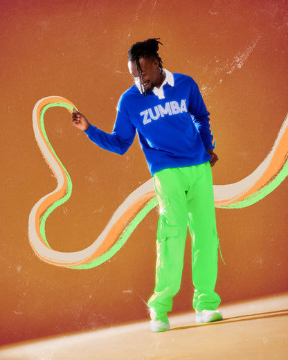 Male model in the neon green tassel cargo pants and a blue long sleeve polo with a white collar and white 'ZUMBA' text, against an orange background with a cream and green swirl.