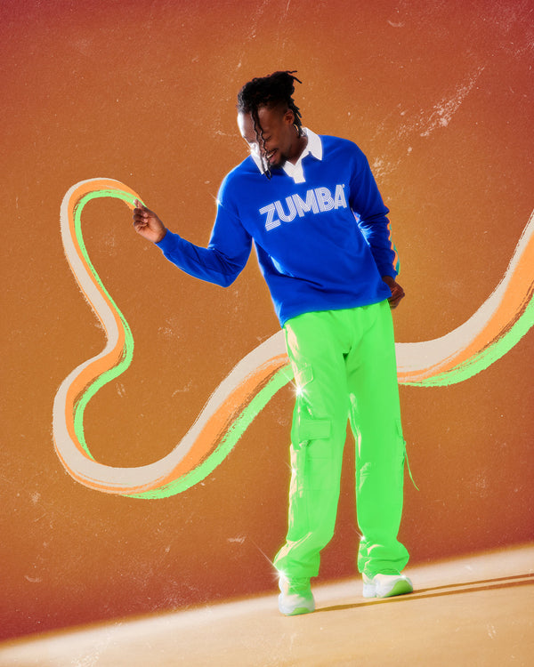 Male model in the neon green tassel cargo pants and a blue long sleeve polo with a white collar and white 'ZUMBA' text, against an orange background with a cream and green swirl.