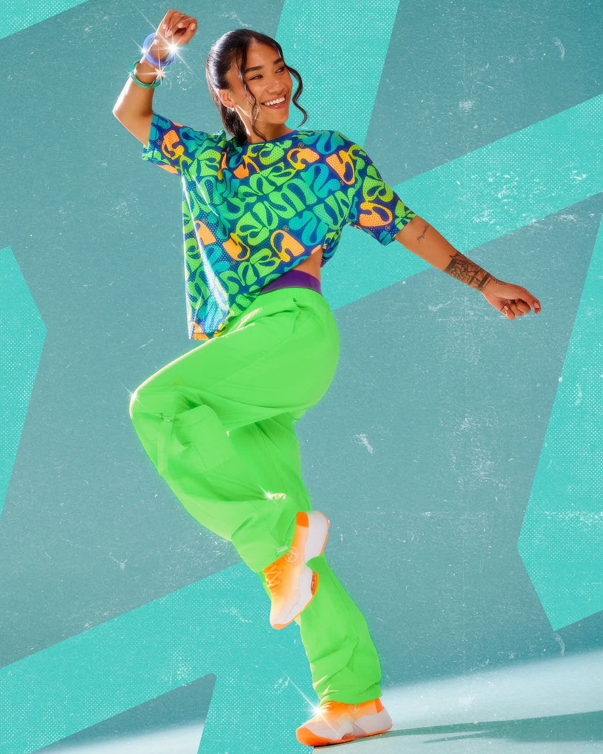 Female model mid-dance in the neon green cargo pants with tassels and a blue warped 'ZUMBA' print mesh top, against an aqua background with star shapes.