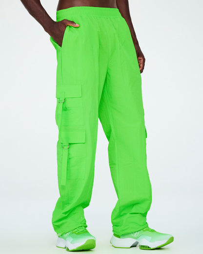 Male model in neon green high-rise cargo pants with an elastic waistband, side cargo pockets with dangling tassels, and drawcords at the hem, one hand in the pocket, paired with neon green shoes.