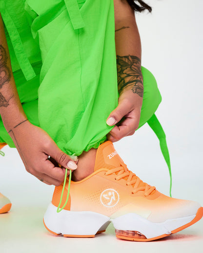 Close-up of hands pulling the bungee drawcord at the hem of the neon green cargo pants to cinch the leg opening above an orange shoe.