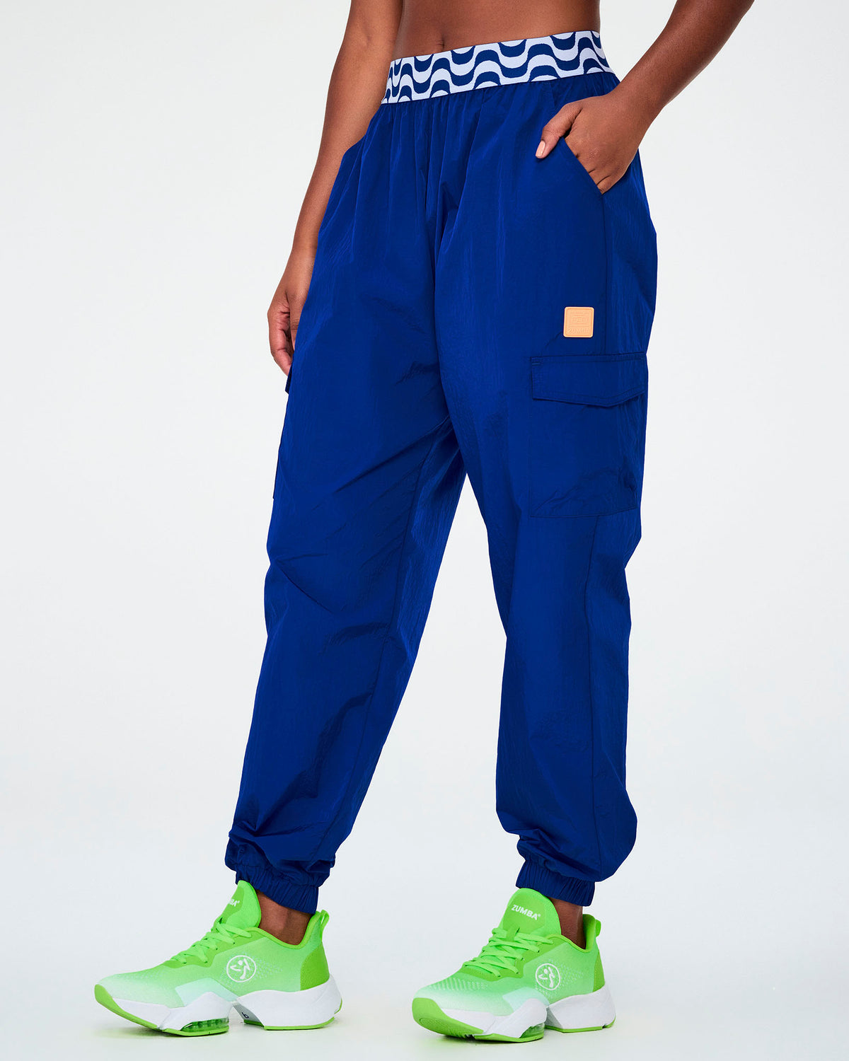 Female model in blue parachute cargo pants with a blue and white wavy print elastic waistband, an orange square tab on the left cargo pocket, and elastic cuffs, one hand in the pocket, paired with neon green shoes.