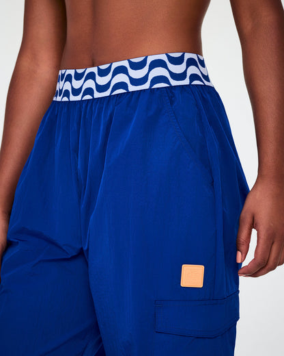 Close-up of the blue parachute cargo pants, showing the blue and white wavy elastic waistband, slanted pocket, and orange square tab on the cargo pocket.