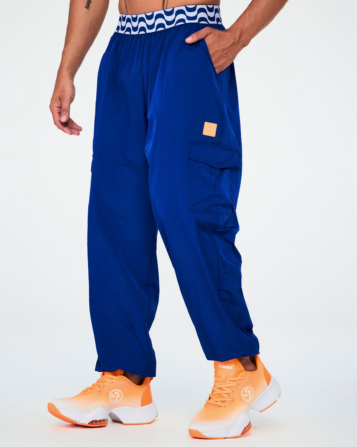 Male model in blue parachute cargo pants with a blue and white wavy print elastic waistband, an orange square tab on the left cargo pocket, and elastic cuffs, one hand in the pocket, paired with orange shoes.