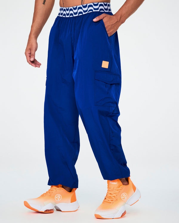 Male model in blue parachute cargo pants with a blue and white wavy print elastic waistband, an orange square tab on the left cargo pocket, and elastic cuffs, one hand in the pocket, paired with orange shoes.