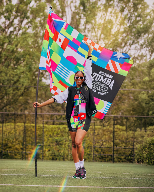 Female model outdoors posing on grass holding a patterned flag with one square in black and white with “United by Zumba” text.