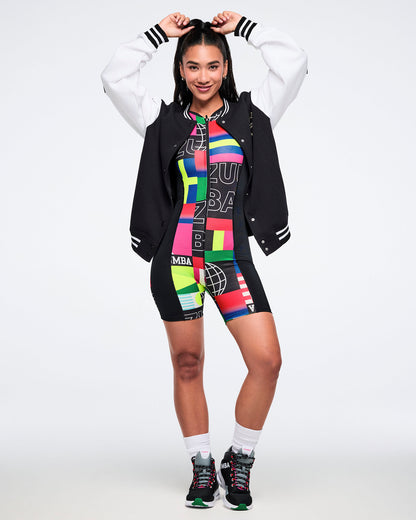 Female model with hands on her head wearing a colorful bodysuit with a black varsity jacket, multicolored shorts, white socks and black Zumba shoes on a white background.