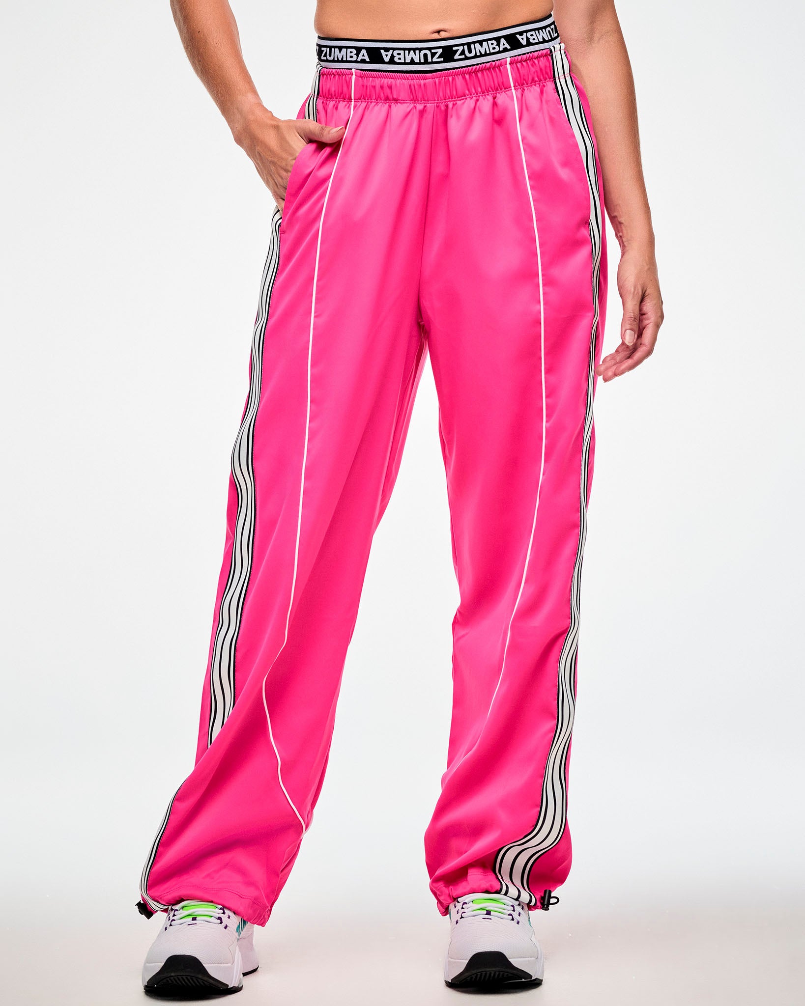 Female model wearing pink track pants with a white and black side stripe and white sneakers, hand in the side pocket.