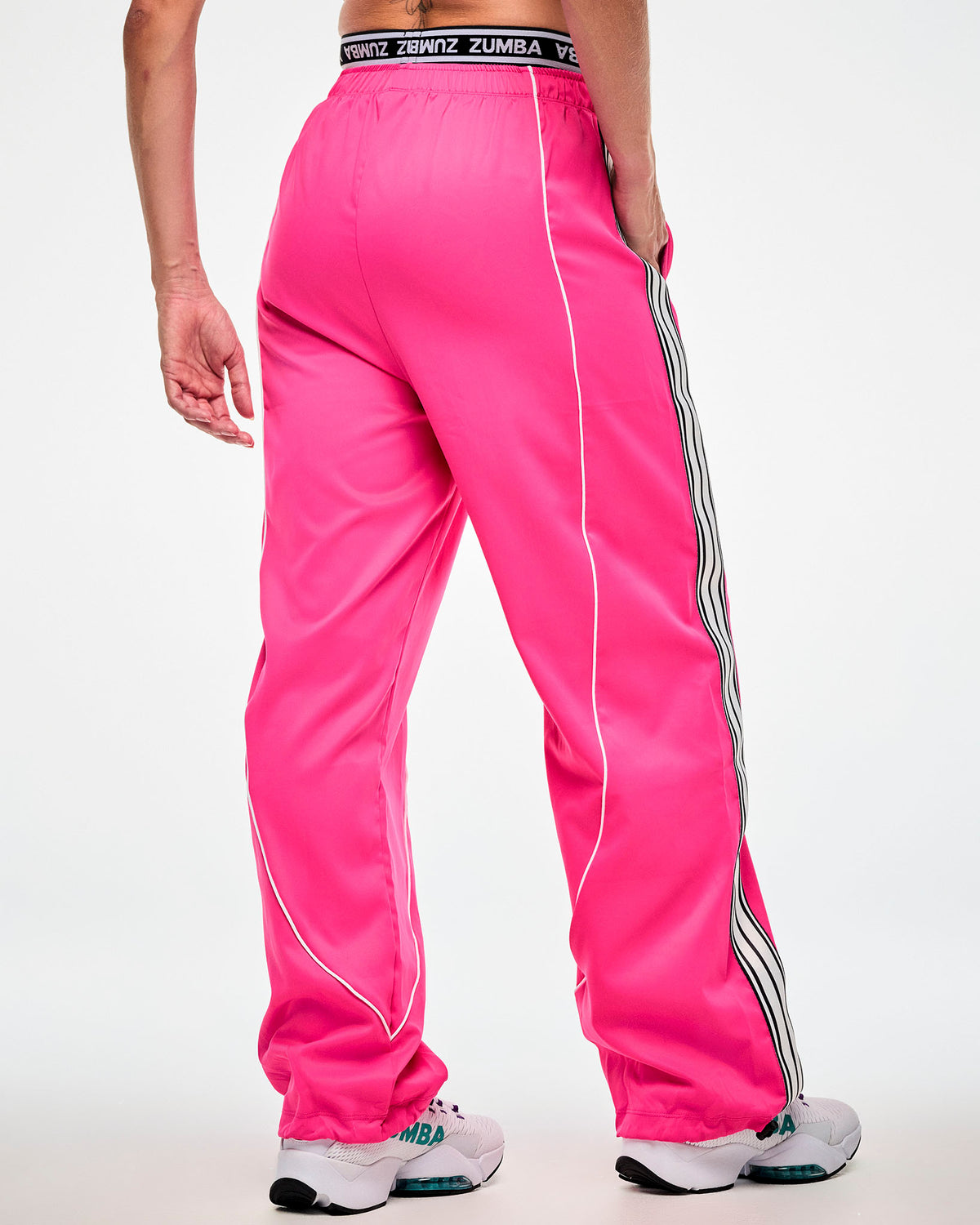 Shown from the back, female model wearing pink track pants with a white and black side stripe and white sneakers, hand in the side pocket.