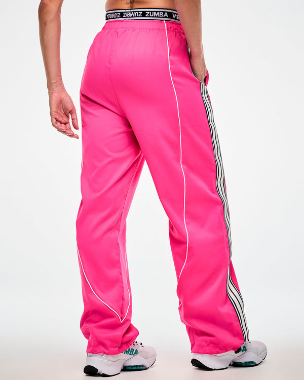 Shown from the back, female model wearing pink track pants with a white and black side stripe and white sneakers, hand in the side pocket.