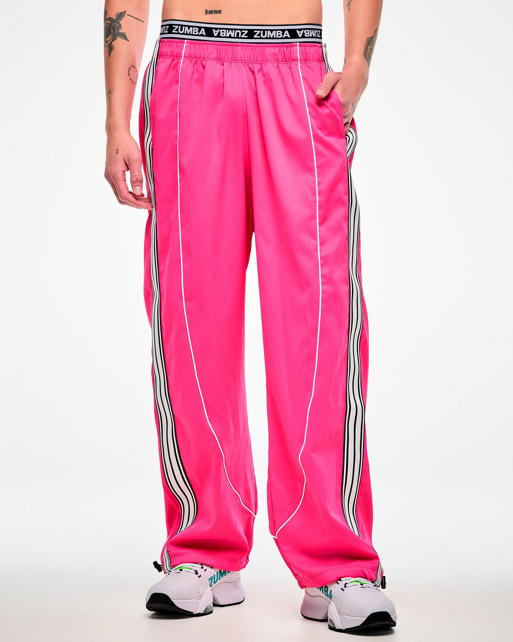 Female model wearing pink track pants with a white and black side stripe and white sneakers, hand in the pocket.