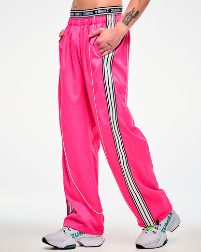 Male model wearing pink track pants with a white and black side stripe and white sneakers, hand in the pocket, side view.