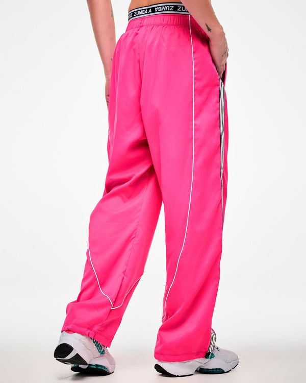 Shown from the back, female model wearing pink track pants with a white and black side stripe and white sneakers, hand in the side pocket.