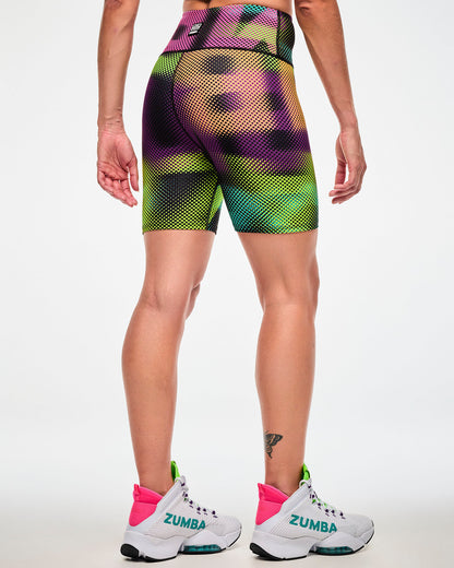 Shown from the back, female model wearing biker shorts with a blurred wordmark print, a Zumba cassette logo on the upper middle waistband and paired with pink sneakers.