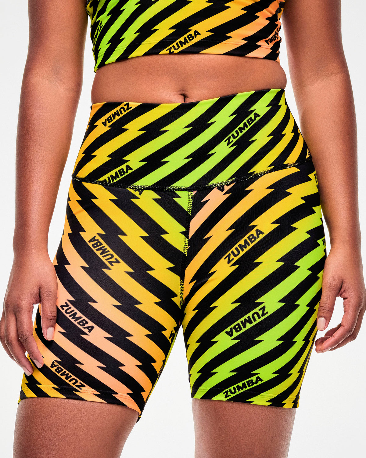 Close-up, side view, of a female model wearing yellow and orange ombre high-waisted biker shorts with a black lightning bolt print and repeated black "ZUMBA" text.