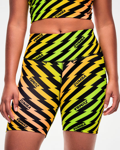 Close-up, side view, of a female model wearing yellow and orange ombre high-waisted biker shorts with a black lightning bolt print and repeated black "ZUMBA" text.