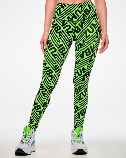 Female model wearing green leggings with a repeated black "ZUMBA" text print and paired with white shoes.