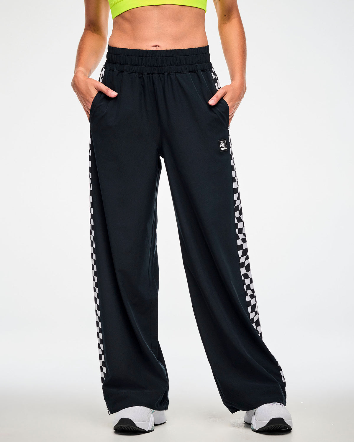 Female model wearing black track pants with a checkerboard side panel and white sneakers, hands tucked in the side pockets.