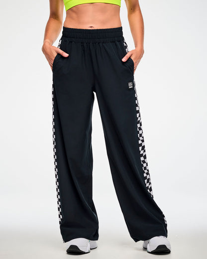 Female model wearing black track pants with a checkerboard side panel and white sneakers, hands tucked in the side pockets.