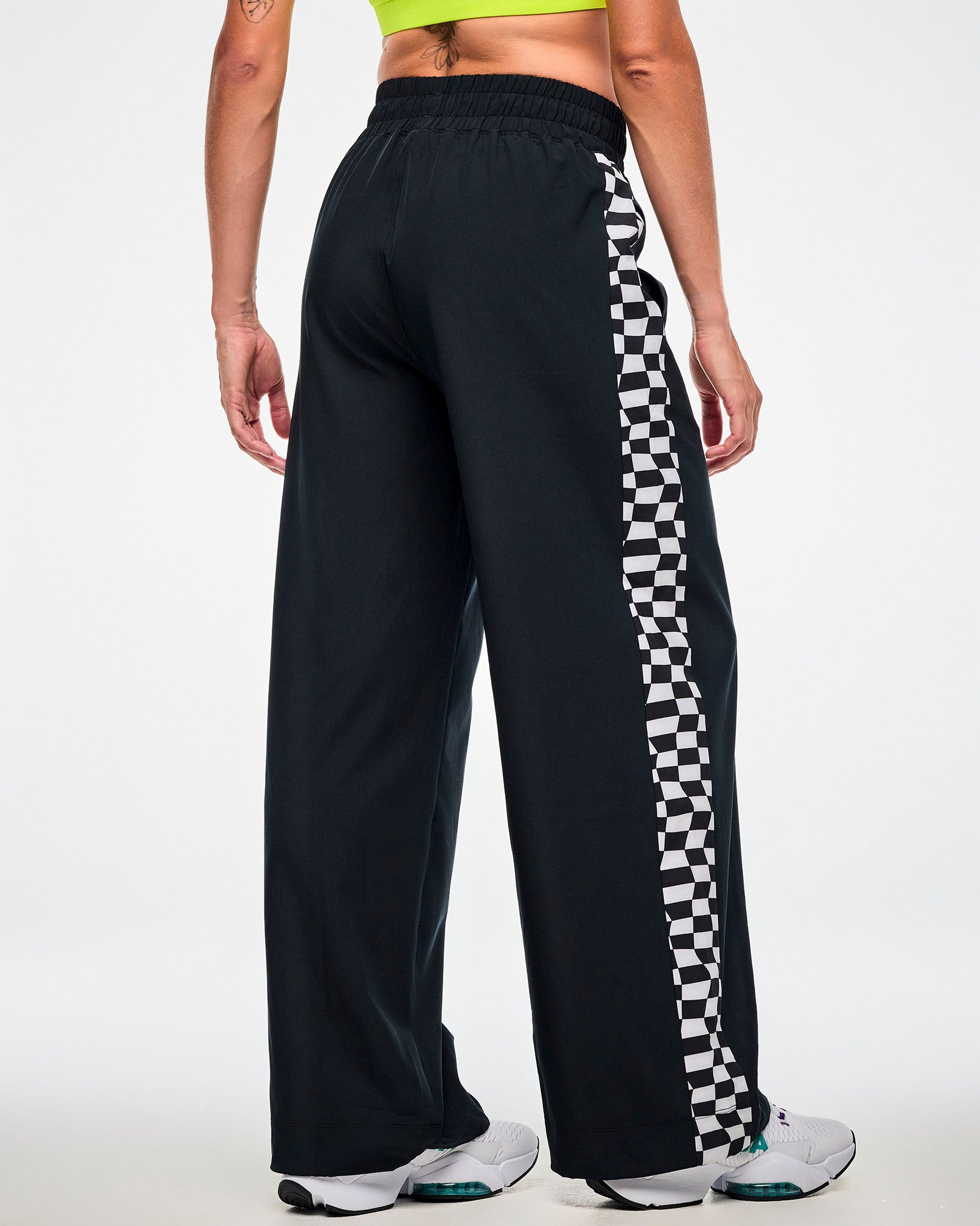 Back view of a female model wearing black track pants with a checkerboard side panel and white sneakers.