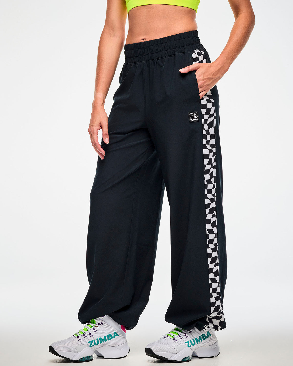 Female model wearing black track pants with a black and white checkerboard side panel, hand in the pocket and paired with white "ZUMBA" sneakers.