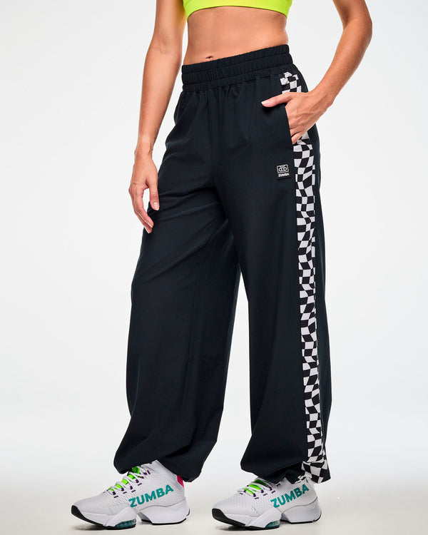 Female model wearing black track pants with a black and white checkerboard side panel, hand in the pocket and paired with white 
